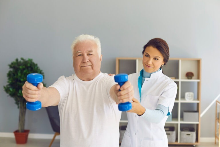 Experienced female doctor supports senior man's arms as he lifts dumbbells in medical office